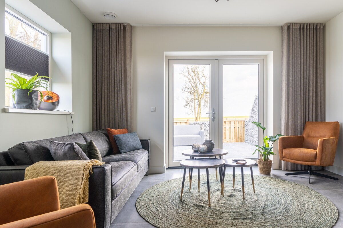 A light-filled living area features a comfortable gray sofa and a rust-colored armchair. A round, textured rug anchors the space, while a wooden coffee table is positioned in the center. Large windows look out onto a private outdoor seating area, framed by flowing curtains.