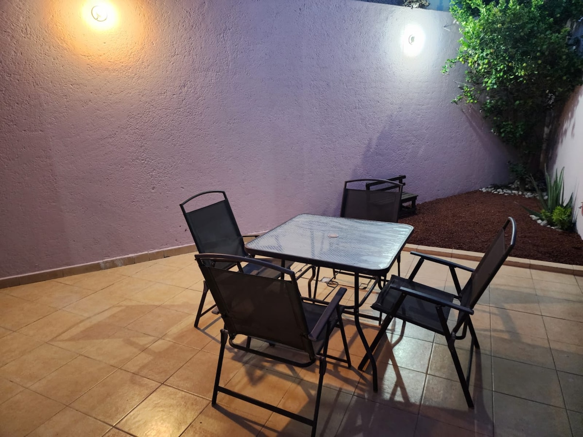 A well-lit outdoor patio area features a glass-top table surrounded by four chairs. The space is bordered by a textured wall with soft lighting, creating a relaxing atmosphere. Decorative plants are positioned along the edges, complementing the stone pathway.