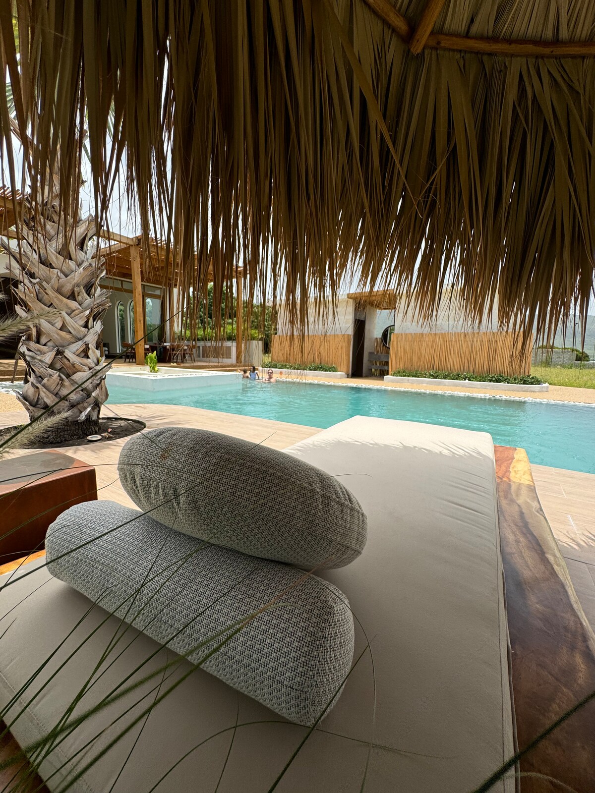 A relaxing poolside scene is presented, featuring a spacious lounging area with large, soft cushions. The water of the pool reflects natural greenery and the thatched roof overhead provides shade, enhancing the tranquil setting.