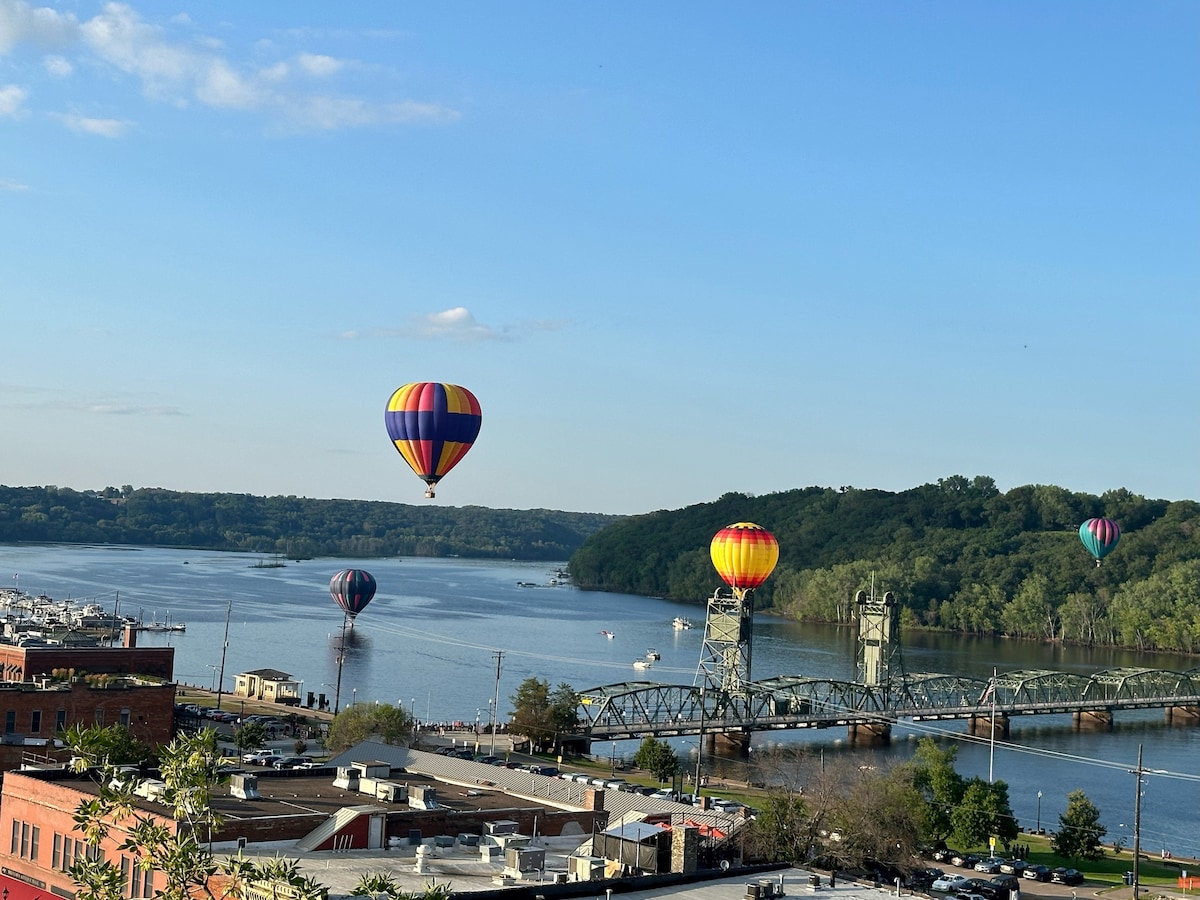 A vibrant scene of colorful hot air balloons floats above the St. Croix River, with the historic Lift Bridge in the foreground. Lush green hillsides frame the landscape, while the calm water reflects the balloons’ hues under a clear blue sky.