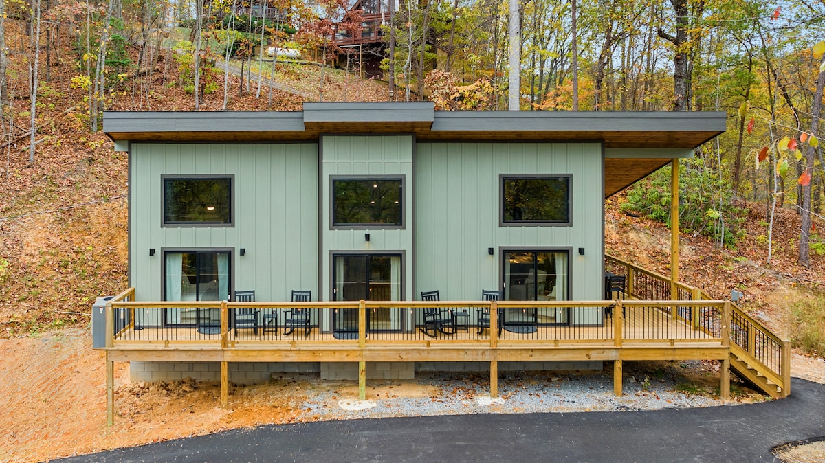 The exterior of the cabin presents a modern design with light green siding and a spacious deck. The front porch features multiple black chairs for seating. Surrounded by trees, the cabin integrates into the natural landscape, offering a welcoming retreat.