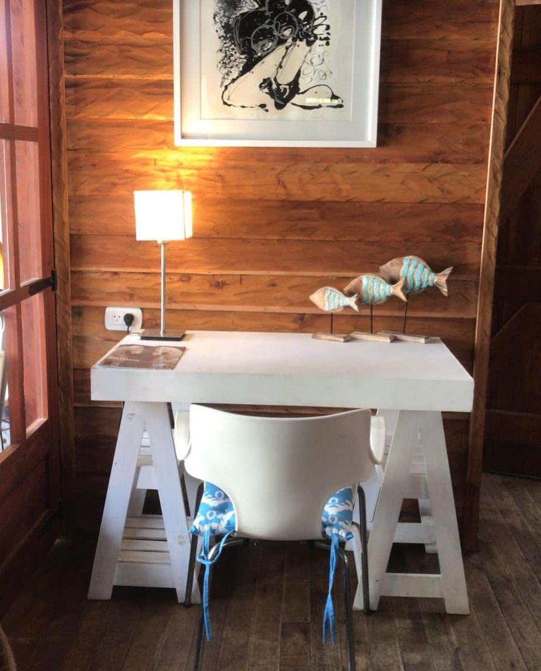 A bright workspace is shown with a minimal white desk and an eye-catching art piece mounted above. A lamp provides soft lighting, while a decorative fish sculpture adds a modern touch. Natural wood walls enhance the room's warmth and inviting atmosphere.