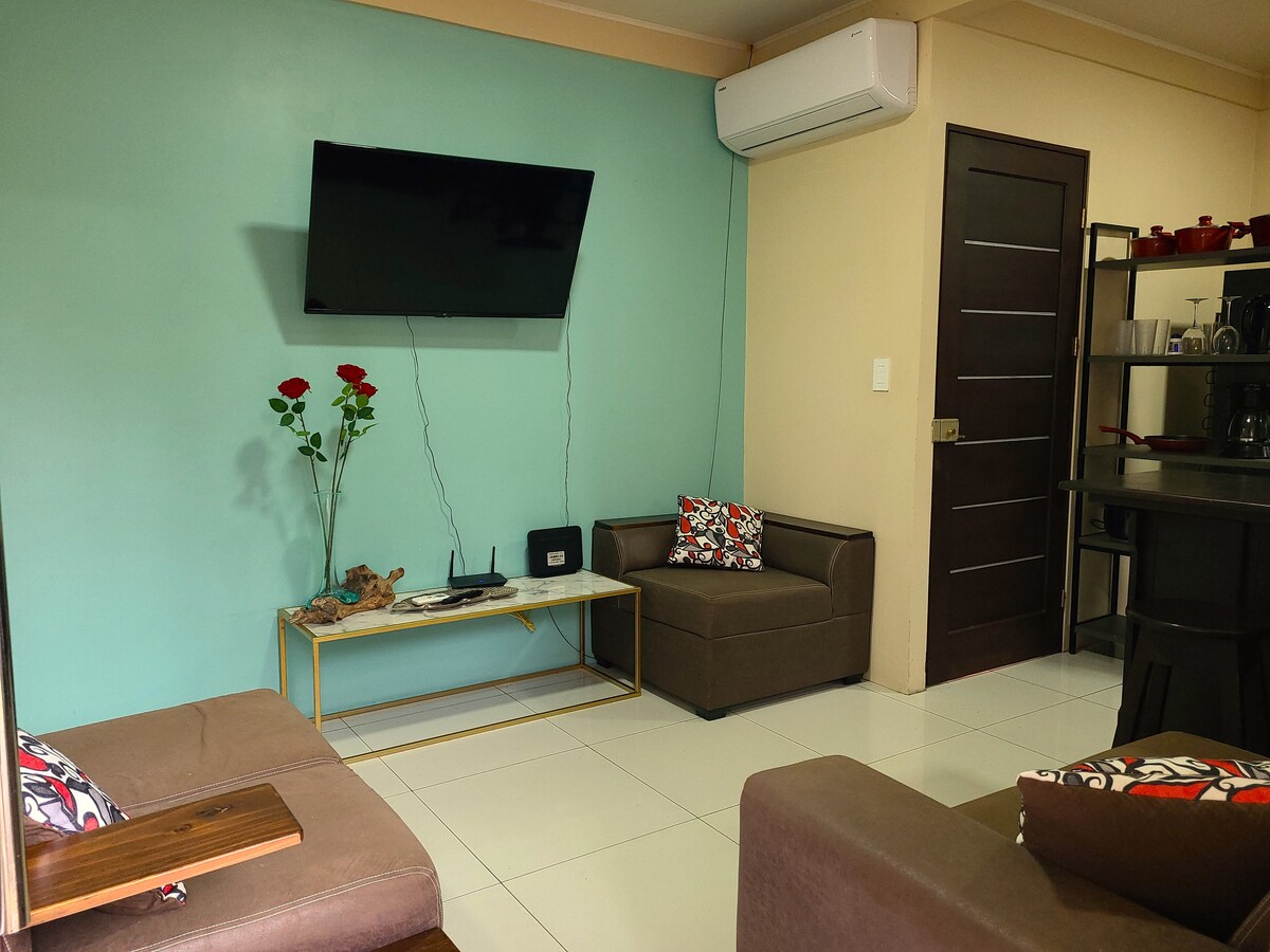A inviting living area features two comfortable sofas adorned with colorful cushions. A flat-screen TV is mounted on a teal wall, while a coffee table sits centrally. Decorative roses are displayed in a vase, adding a touch of elegance to the space.