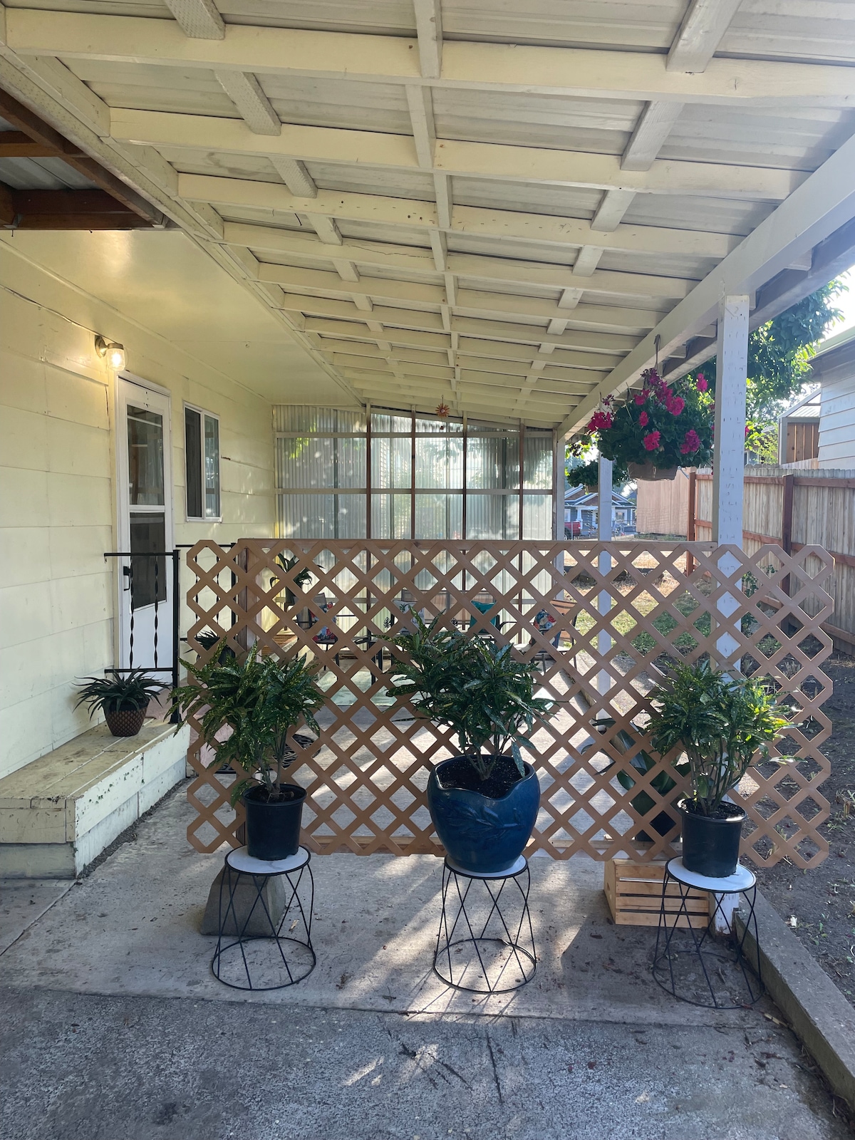 A covered patio area features a light-colored lattice partition with potted plants on small tables. The entrance to the guest house is visible, along with a wall of greenery in the background. Sunlight filters through the space, creating a welcoming environment.
