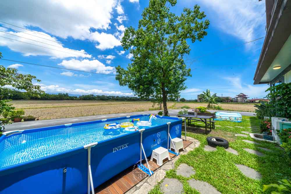An outdoor area is showcased with an above-ground pool surrounded by lush greenery and a tree. Nearby, a barbecue area is set up with a table and seating. A clear sky with scattered clouds creates a bright, inviting atmosphere.