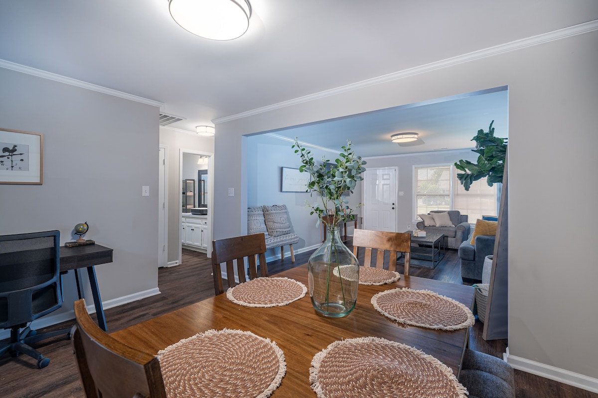 A dining area is displayed with a wooden table surrounded by four chairs. A large glass vase filled with greenery serves as the centerpiece. Natural light is visible through a doorway leading to a living space, enhancing the open feel of the room.