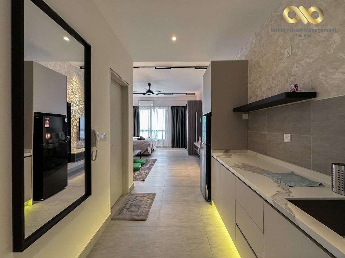 A modern kitchen area is visible with sleek cabinetry and a large mirror reflecting the space. The layout includes a refrigerator and countertop, leading towards a bright living area. Natural light filters in from a window, enhancing the ambiance of the open space.