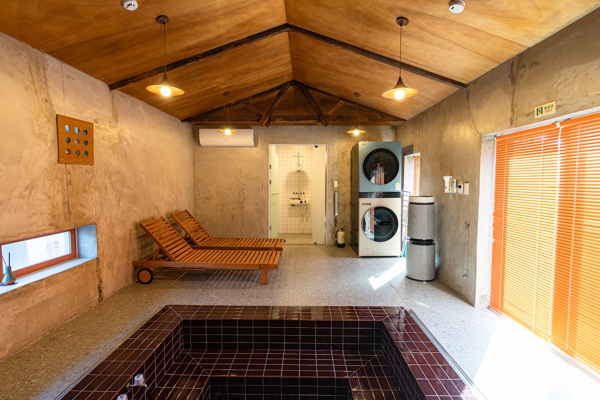 A spacious relaxation area features wooden lounge chairs arranged beside a built-in hot tub. The room is illuminated by pendant lights, while a shower area is enclosed in glass. A set of laundry machines is visible in the background, and large orange-blind windows provide natural light.