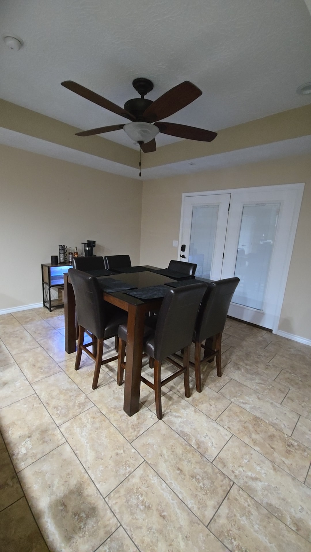 A dining area features a wooden table surrounded by six black leather chairs. A ceiling fan is mounted above, and light-colored walls complement the tile flooring. Double glass doors lead to another space, while a small beverage station is visible in the corner.