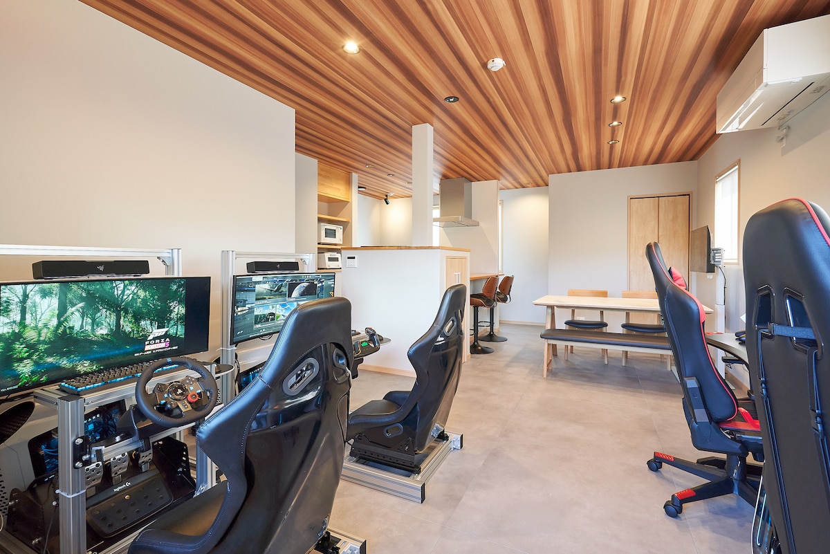 A spacious gaming area features high-end gaming PCs and driving simulators, surrounded by a sleek design. Wood-paneled ceilings enhance the modern aesthetic, with a dining table and chairs visible in the background, providing a versatile space for relaxation and gaming.