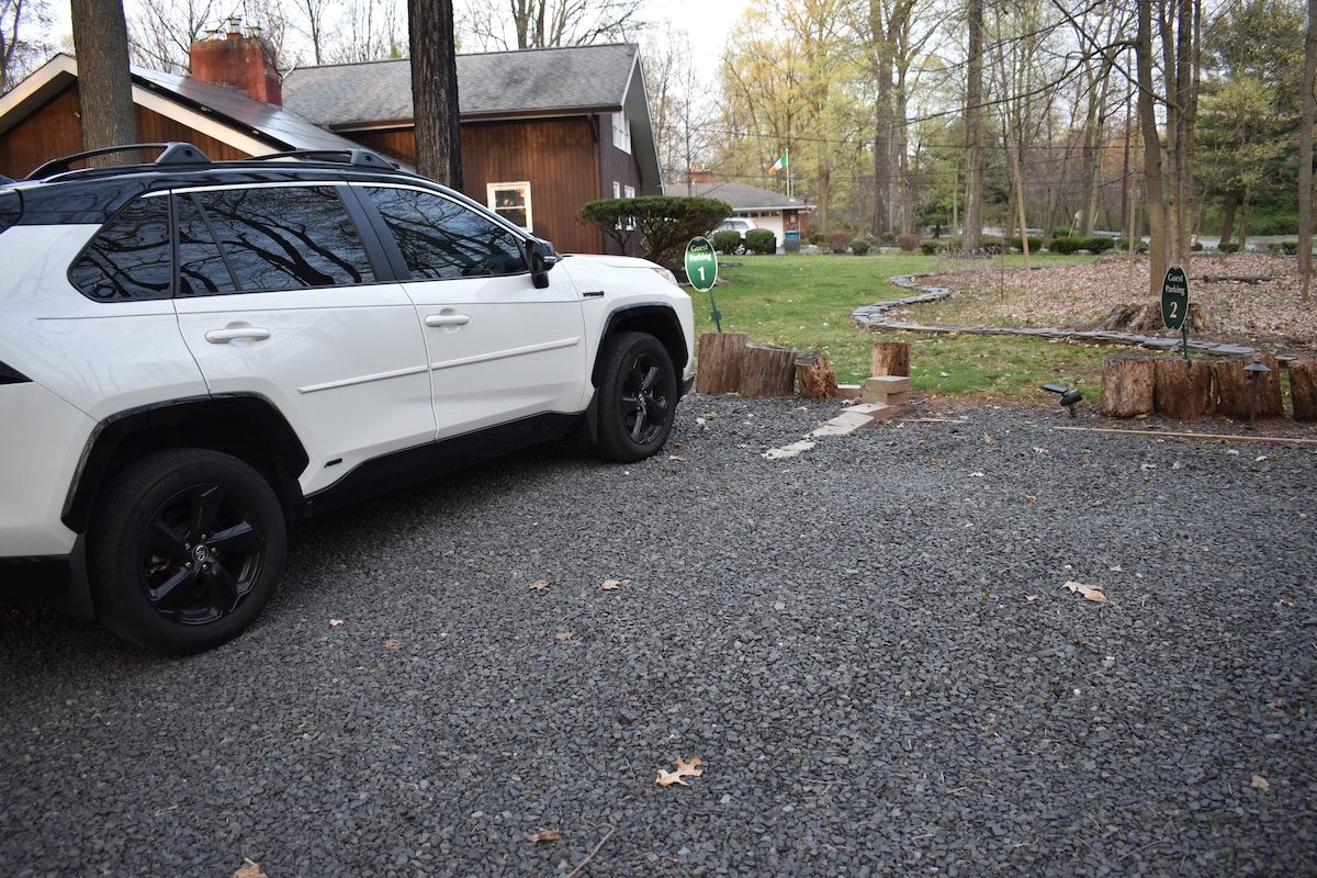 A gravel parking area is shown, featuring a white vehicle parked beside a tree stump. A pathway leads toward a grassy yard, surrounded by trees and landscaped with stones. The welcoming ambiance is highlighted by the natural surroundings.