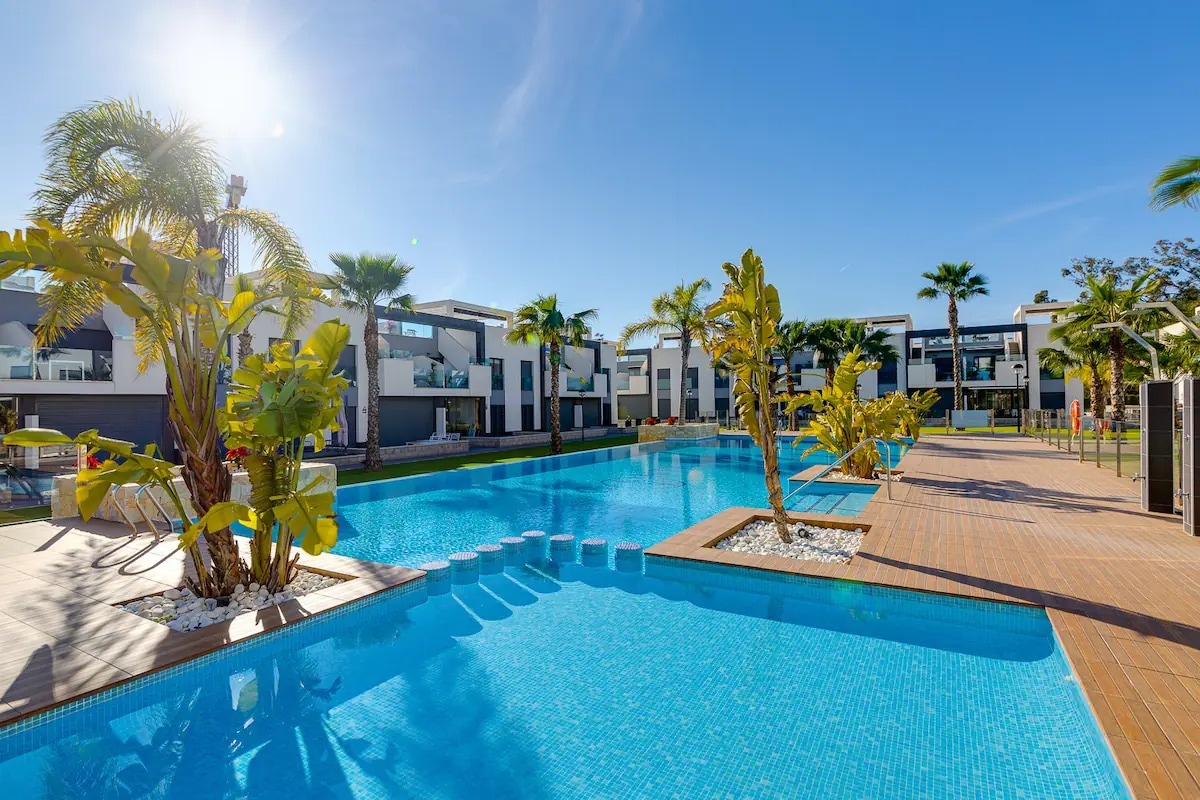 A serene outdoor area features a large swimming pool surrounded by palm trees and lush greenery. The clear blue water reflects the sunlight, while neighboring buildings provide a modern backdrop. Poolside lounging chairs are positioned along the wooden deck, inviting relaxation.