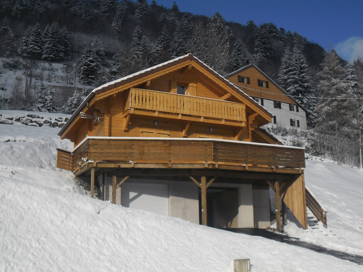A wooden chalet with a spacious balcony wraps around its upper level, offering sweeping views of the surrounding snowy landscape. The lower level is accessible from the ground, and the structure is set against a backdrop of evergreen trees and a clear blue sky.