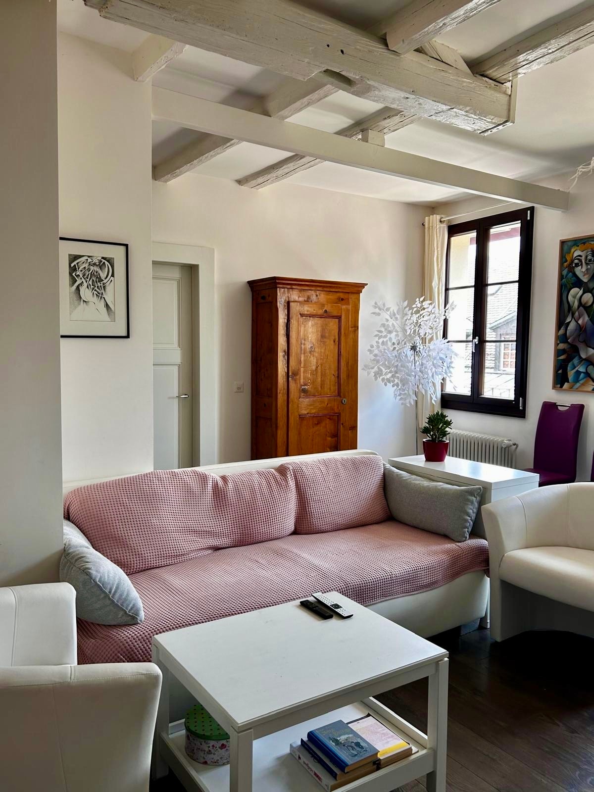 A cozy living area features a light-colored sofa with pink cushions and a white armchair. A small white coffee table holds a book and a remote. In the background, a wooden cabinet and a potted plant add character, while natural light streams through the window with open shutters.