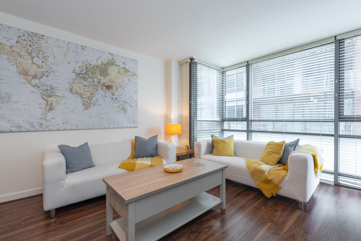 A bright living area features large windows with adjustable blinds, allowing natural light to fill the space. Soft white sofas, adorned with decorative cushions, offer comfortable seating. A wooden coffee table sits centrally, accompanied by a wall-mounted world map, providing a modern and inviting atmosphere.