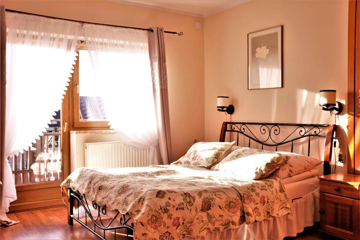 A well-lit bedroom features a spacious bed adorned with a floral patterned quilt. Sheer curtains gently frame the window, allowing natural light to fill the room. A minimalist wall decor piece adds a touch of elegance, while soft wall sconces provide a warm glow.