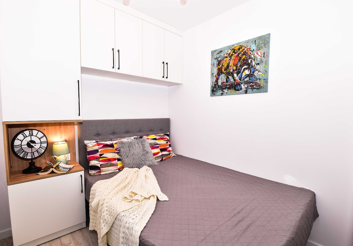 A compact sleeping area features a spacious bed adorned with textured bedding. Colorful throw pillows and a soft blanket add comfort. A framed art piece hangs above the bed, while a bedside table holds a clock and lamp, complementing the modern cabinetry.