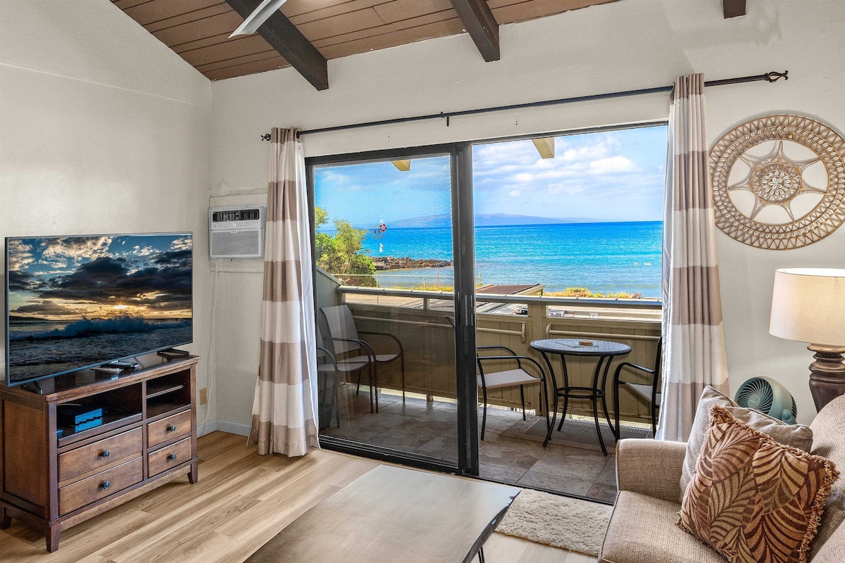 The living area features large sliding glass doors providing a view of the ocean and the outdoor lanai. A flat-screen TV is positioned on a wooden console, complemented by a cozy sofa. Light drapes frame the entrance, enhancing the bright ambiance.