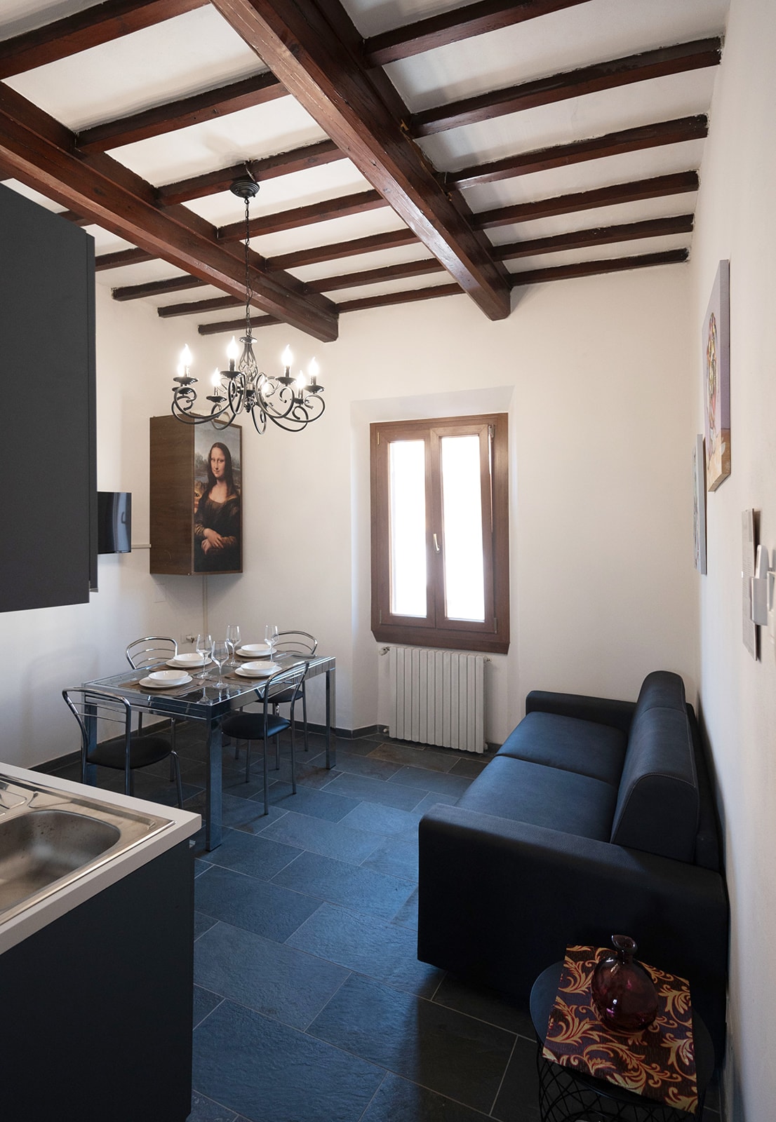 A cozy living area features a dark sofa opposite a glass dining table set for four. Natural light enters through a window with wooden frames, illuminating the space. Exposed wooden beams line the ceiling, while a chandelier adds a touch of elegance.