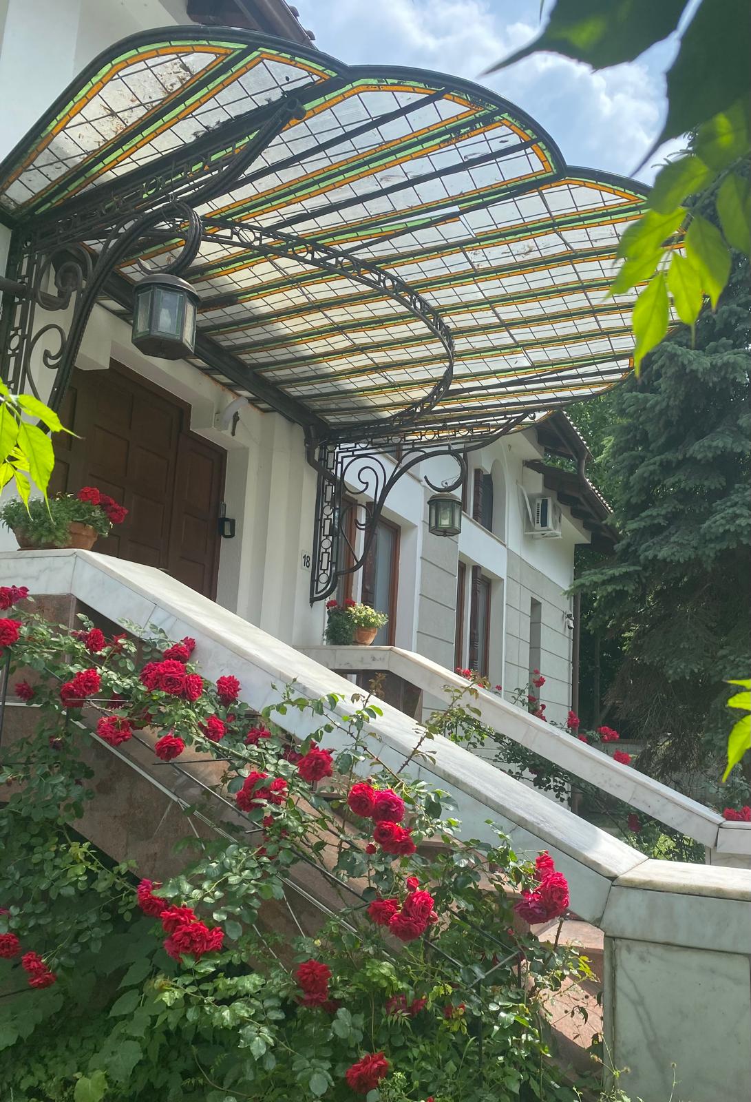 An elegant entrance features a curved glass awning supported by ornate metalwork. Lush red roses climb the stairway, framing the entrance with vibrant blooms. The front door, made of rich wood, presents a welcoming entry to the space.
