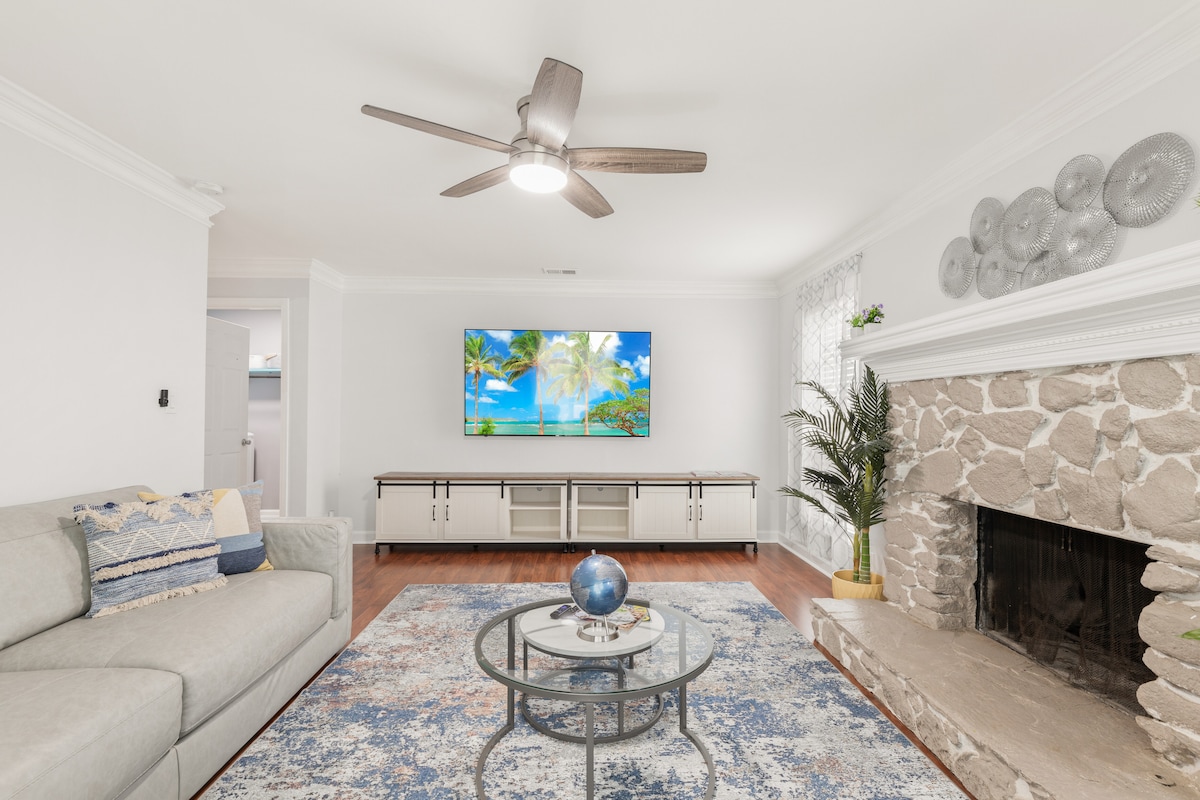A spacious living room is highlighted by a comfortable light grey couch and a round glass coffee table. A large TV is mounted on the wall, and a stone fireplace adds a natural element. Decor includes a decorative plant and a vibrant area rug.