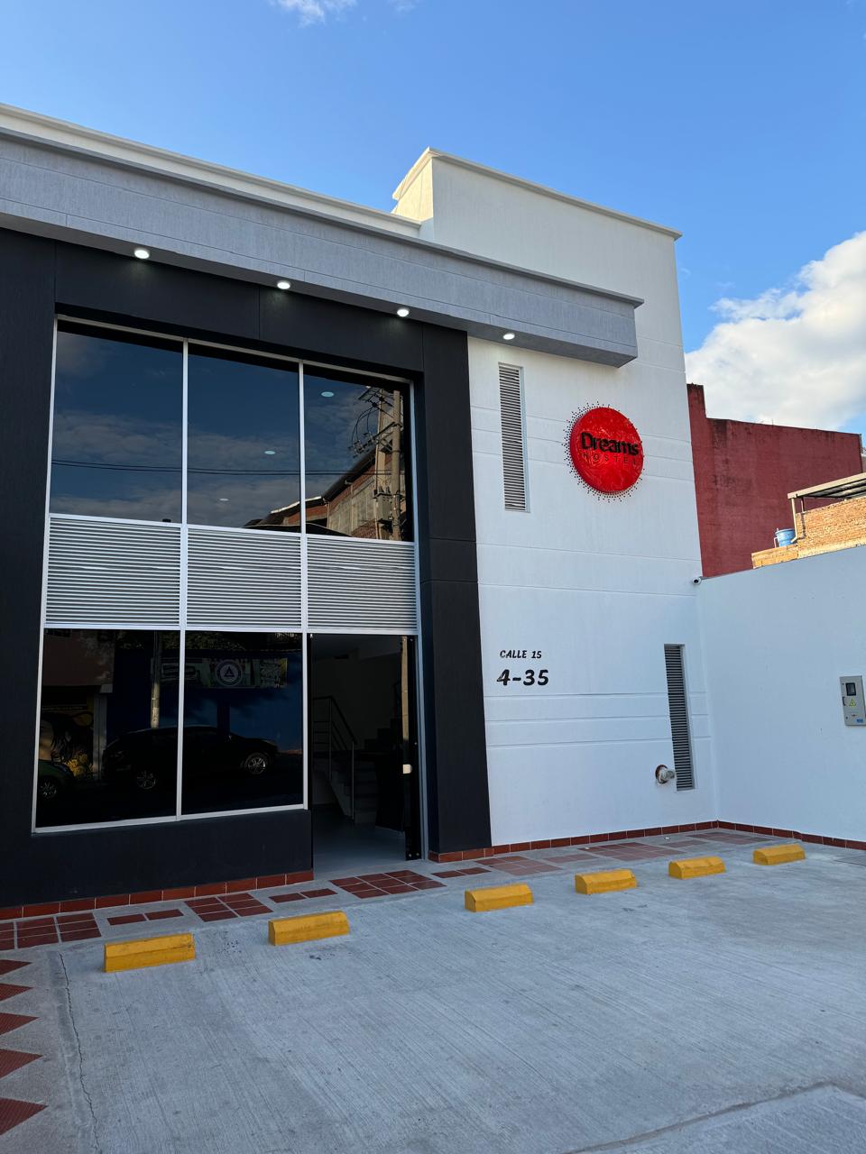 The exterior of a modern building is presented, featuring a large glass front that allows natural light to enter. The structure is complemented by a minimalistic design in black and white, with clear signage indicating the address. A paved area with designated parking spaces is visible in the foreground.