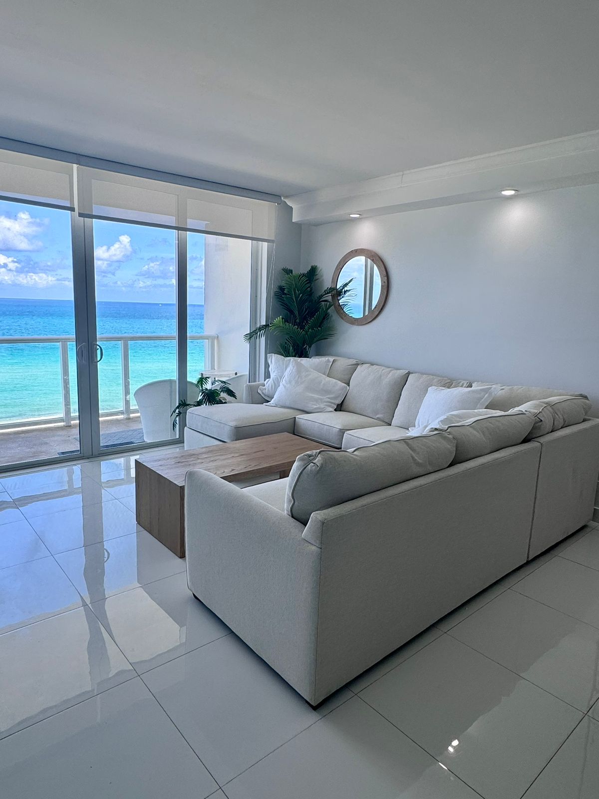 A spacious living area features a light-colored sectional sofa complemented by a wooden coffee table. Large glass doors offer a view of the ocean, allowing natural light to fill the room. A round mirror and potted plant add subtle details to the modern decor.