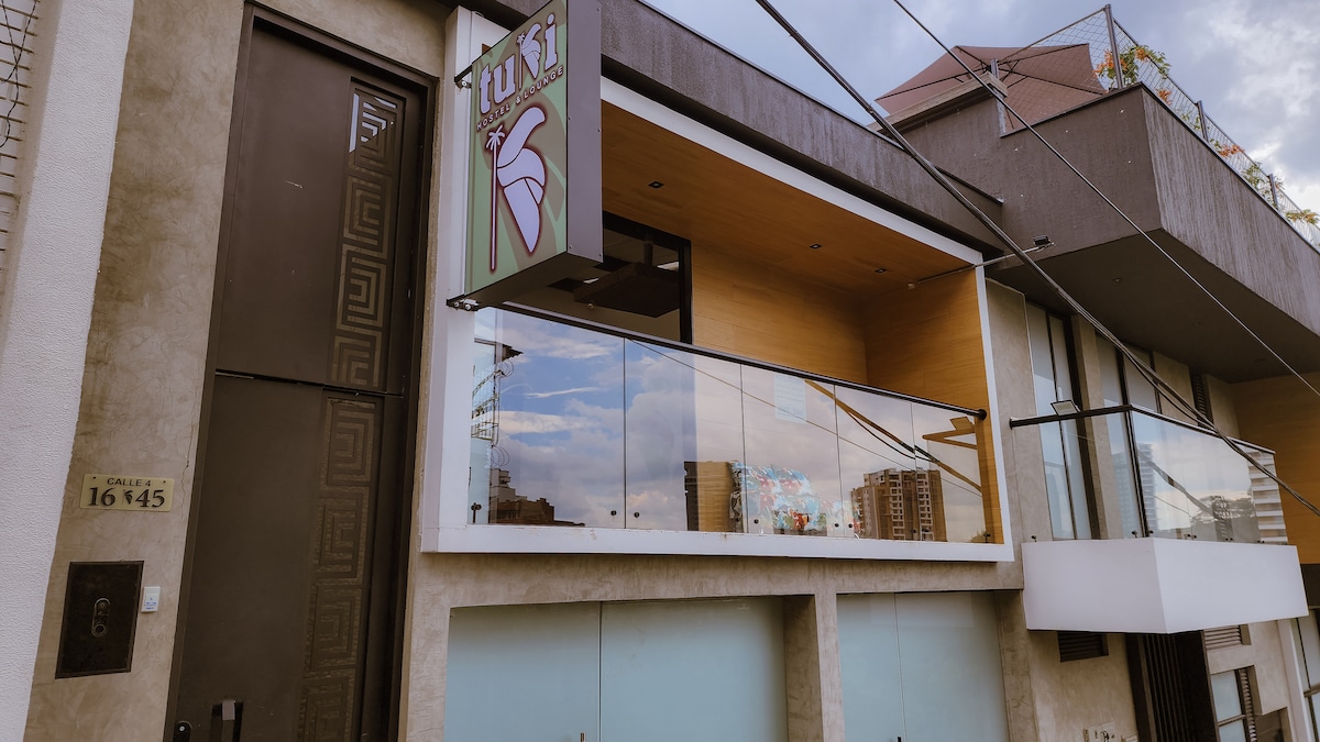 The exterior of Tuki Hostel is displayed, showcasing a modern façade with a large balcony and decorative signage. Large windows reflect the surrounding cityscape, and the entrance is framed by a minimalist design that merges contemporary aesthetics with a welcoming atmosphere.