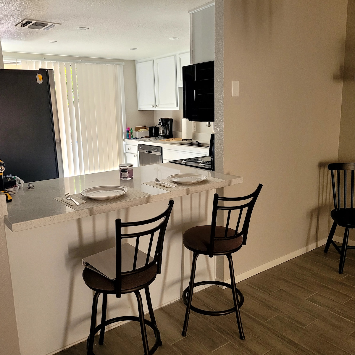 A modern kitchen area features a polished countertop with two high-backed stools. An open space behind includes a black refrigerator and an organized kitchen setup with white cabinetry. Vertical blinds allow natural light to filter in, highlighting the neutral-colored walls and tiled flooring.