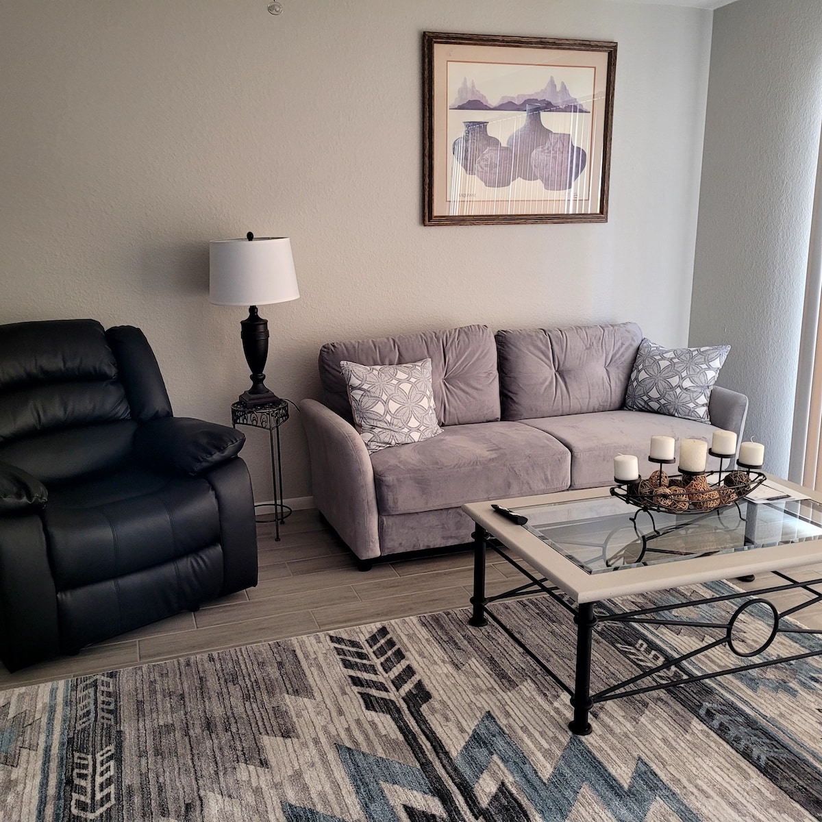 A comfortable living area is showcased, featuring a gray sofa adorned with decorative pillows, alongside a black reclining chair. A coffee table with a glass top is positioned in front, topped with a candle arrangement. A framed piece of art is displayed on the wall.