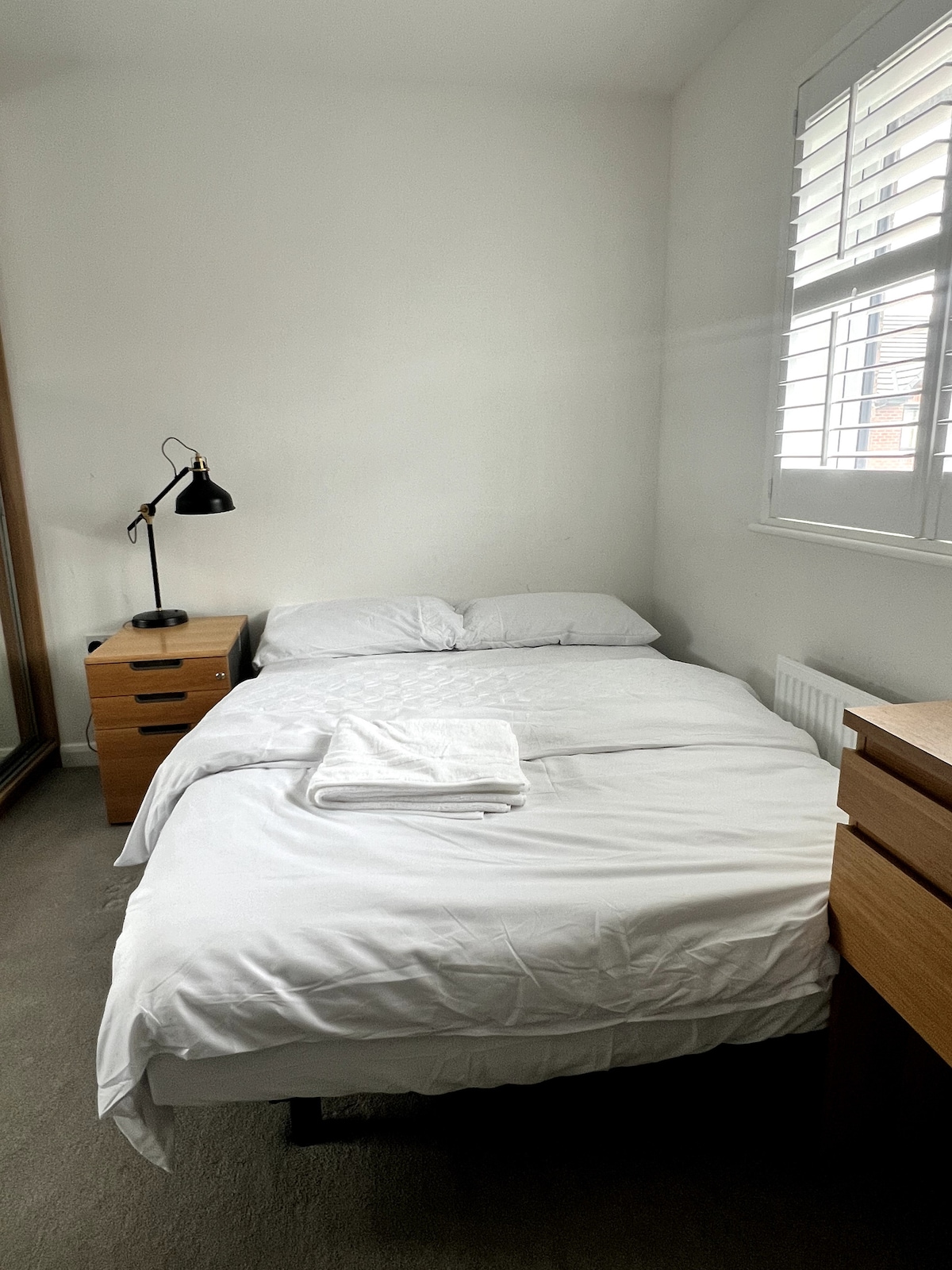 A private bedroom features a spacious bed dressed in white linens. A bedside table is positioned beside the bed, holding a black lamp. Natural light enters through a window with plantation shutters, brightening the neutral-colored walls and carpeting.