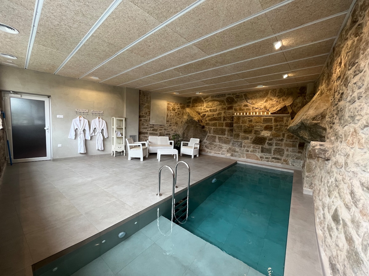 An indoor relaxation area features a turquoise pool with a ladder for easy access. Surrounding the pool, comfortable seating is arranged. Exposed stone walls add a natural element, while bathrobes hang on hooks, indicating a spa-like atmosphere.