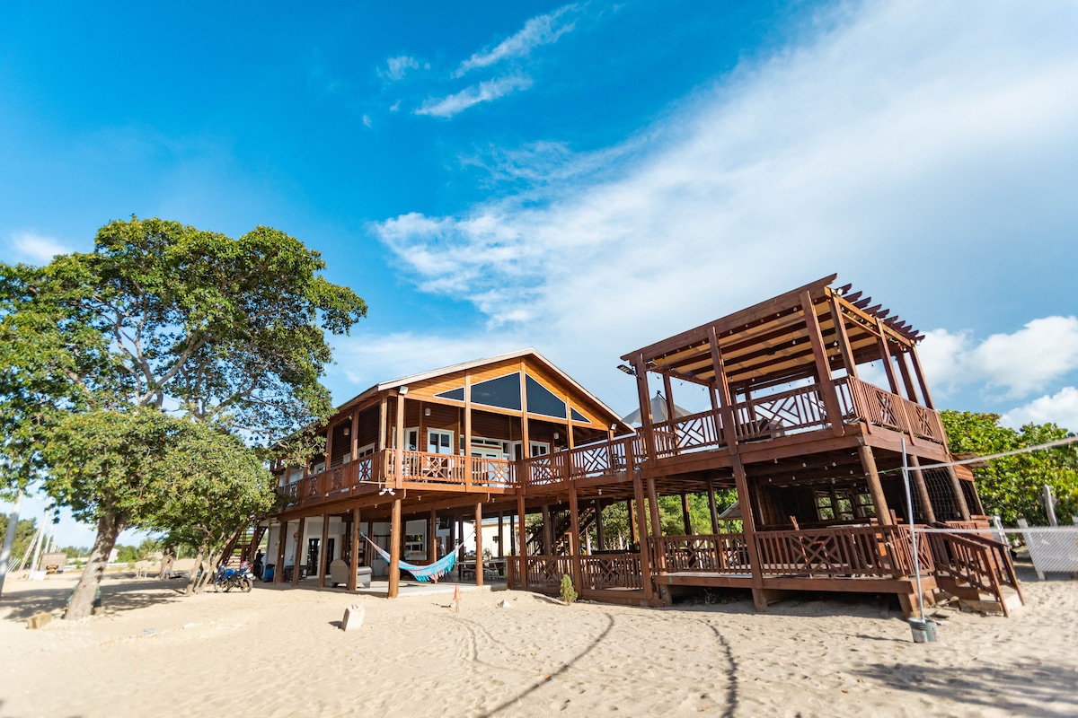 The resort features a large wooden structure with multiple levels, set against a clear blue sky. A spacious deck extends outward, offering seating areas and hammocks. The sandy area surrounding the building invites relaxation, with lush greenery adding to the natural setting.