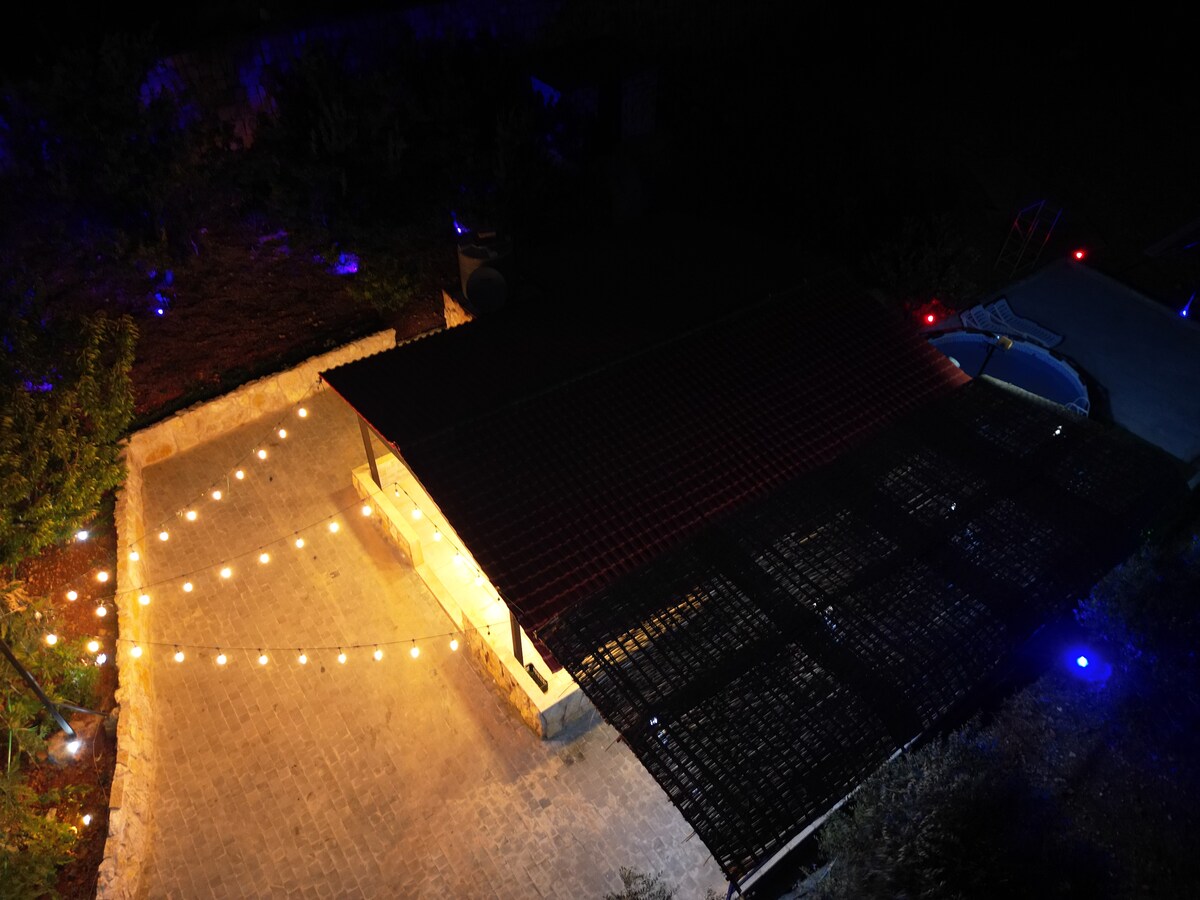 An outdoor area is illuminated by string lights, creating a warm ambiance. The structure features a roof with a slatted design, and a smooth stone surface leads to various outdoor spaces. Soft blue lighting adds a calming touch to the surroundings.