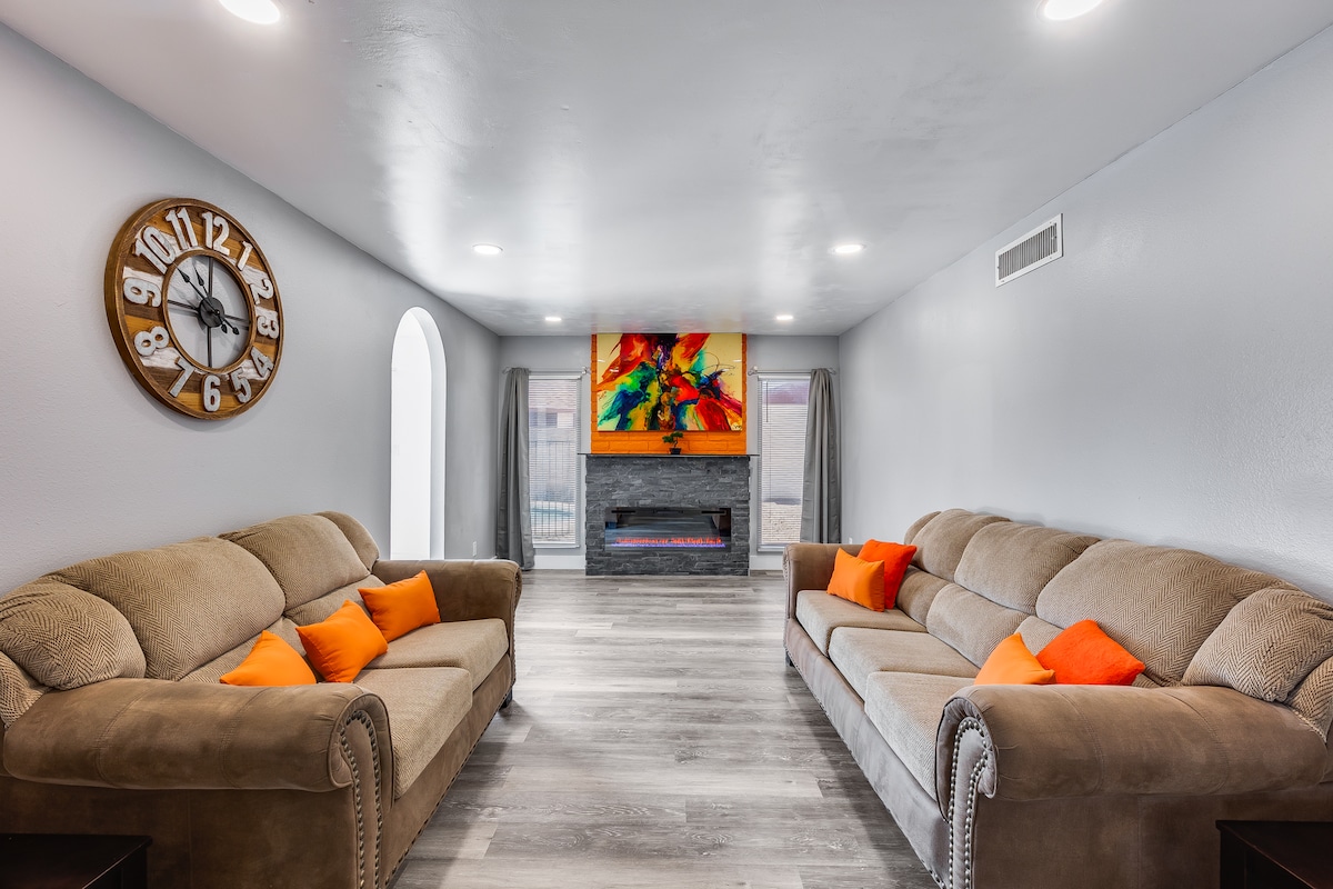 The living area features two spacious sofas adorned with orange accent pillows, offering comfort for gatherings. A large wall-mounted television is situated above a modern fireplace, with vivid artwork displayed above. Natural light enters through a nearby arched window, enhancing the open ambiance.