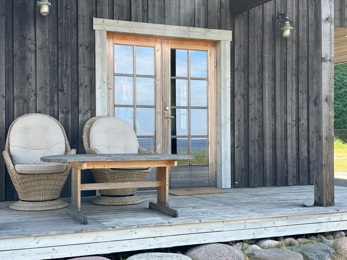 A wooden terrace features two circular, cushioned chairs and a wooden table, creating a relaxed seating area. Large glass doors lead into the sauna house, providing a view of the surrounding landscape and sea beyond.
