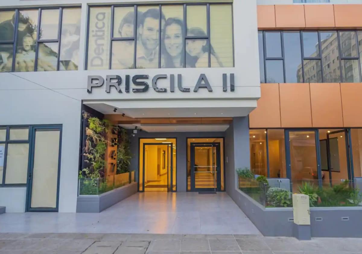 The entrance to the building features a modern facade with large glass windows and a welcoming door. The name 'PRISCILA II' is prominently displayed above the entrance, while plants are arranged thoughtfully along the pathway, creating an accessible and inviting approach.