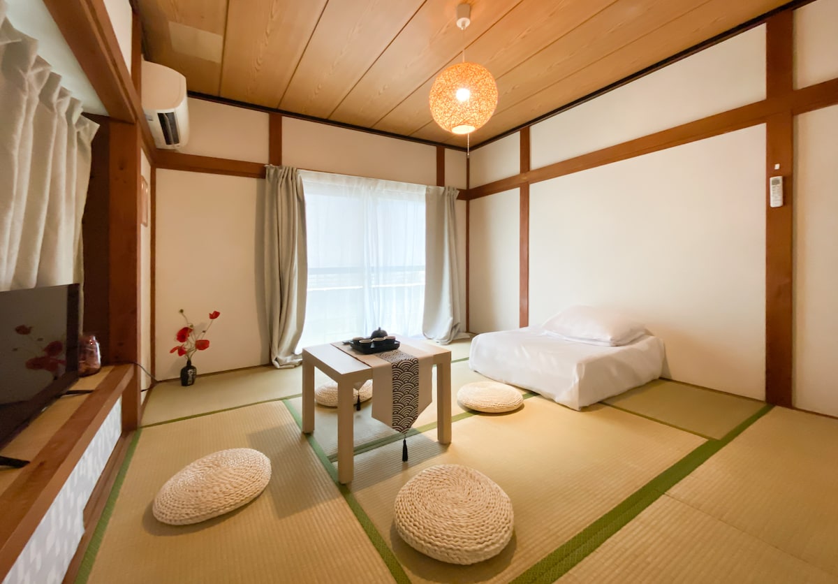 A traditional tatami room features wooden beams and a soft natural light filtering through sheer curtains. Centered is a low table with plush round cushions arranged around it. A single bed is neatly made in one corner, contributing to a serene atmosphere.