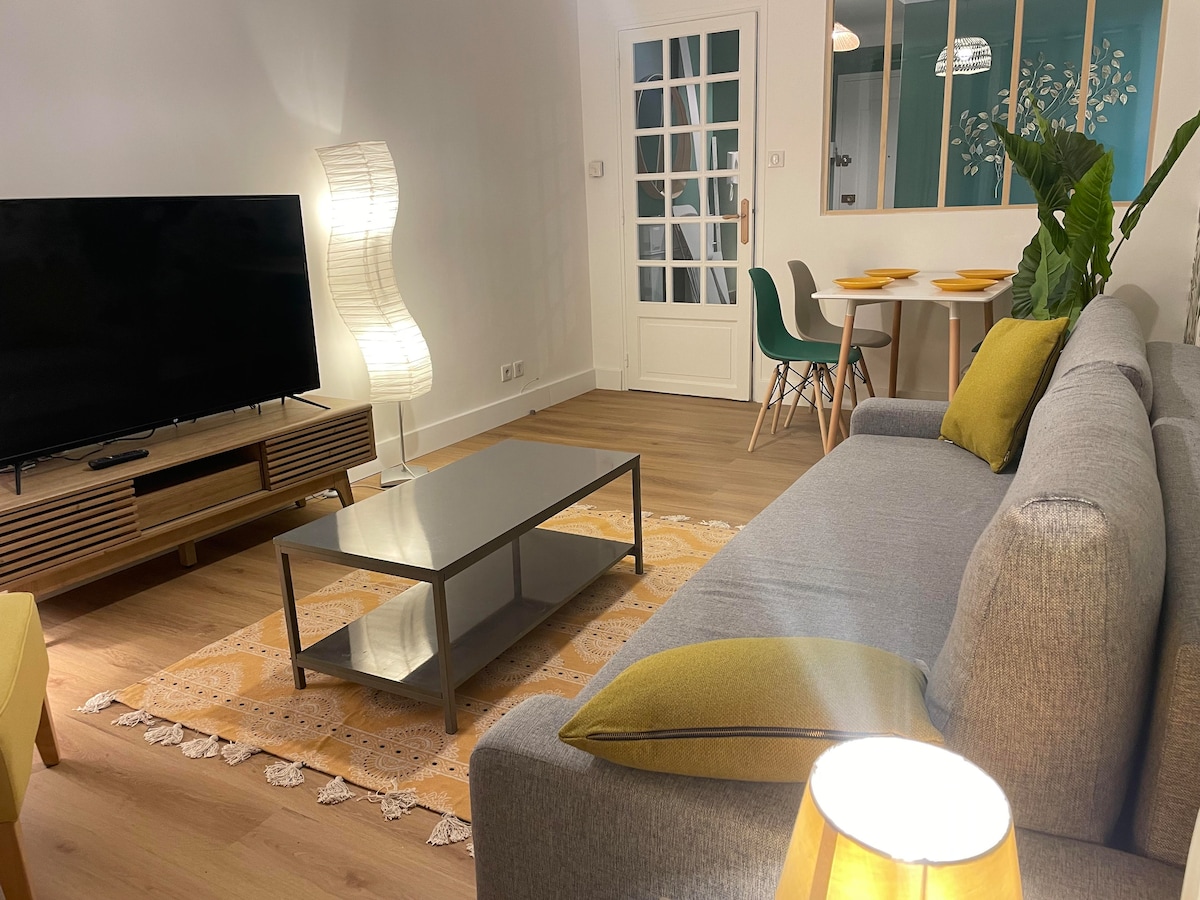 A cozy living area features a plush gray sofa adorned with bright green cushions. In front, a sleek coffee table rests on a decorative rug, while a television is positioned on a low console. A dining table is visible in the background, accompanied by modern chairs.