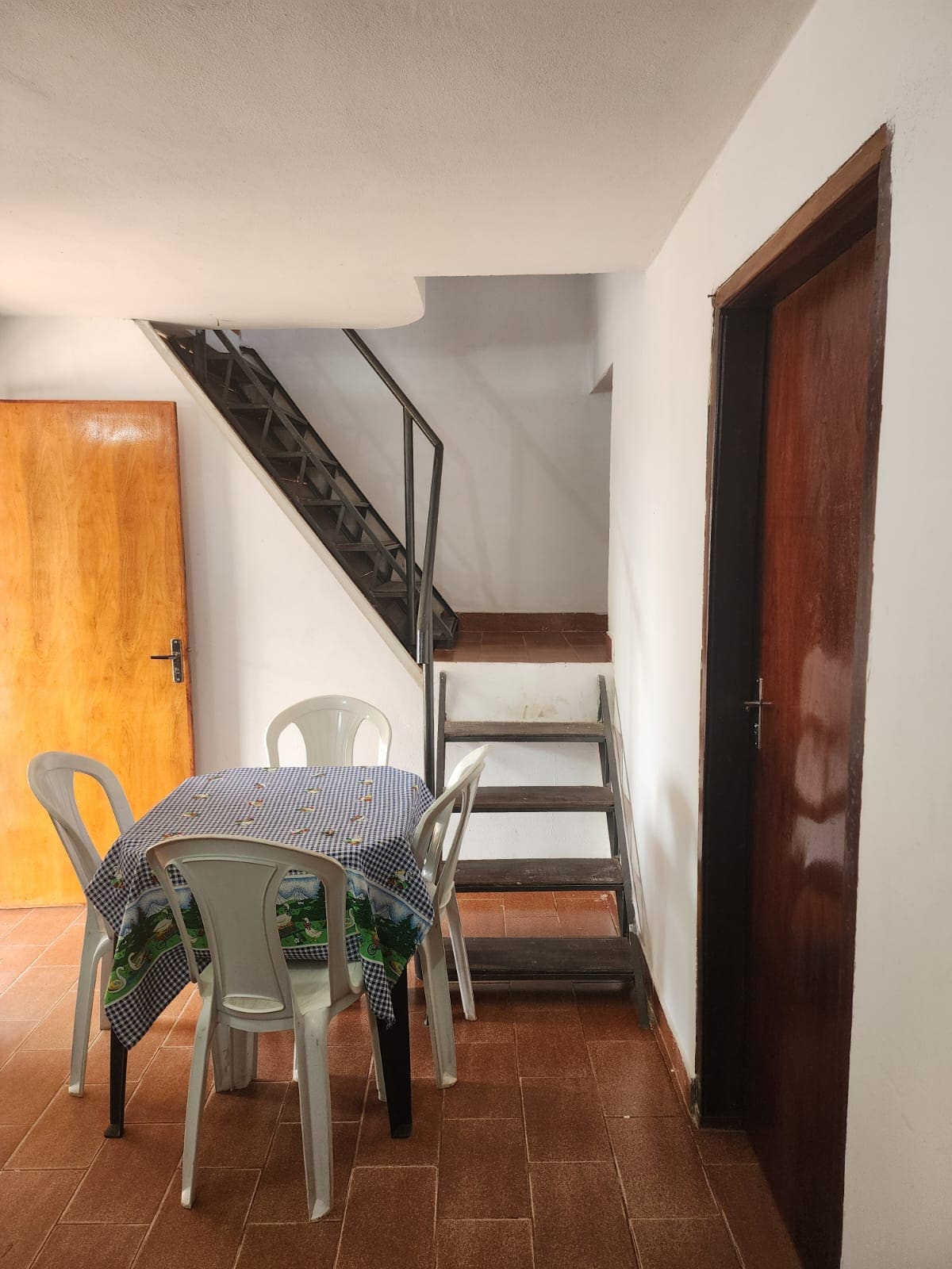 A simple dining area displays a checkered tablecloth covering a round table with four white chairs. A wooden door leads outside, and a staircase is visible in the background, with a metal railing providing access to the upper level.