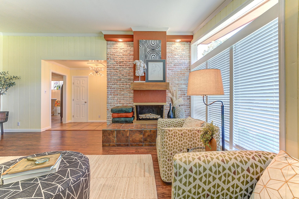 The living room features a brick fireplace as a focal point, flanked by large windows with horizontal blinds allowing natural light. Comfortable seating includes patterned armchairs and a textured area rug, creating a cozy atmosphere. A small lamp adds warmth to the space.