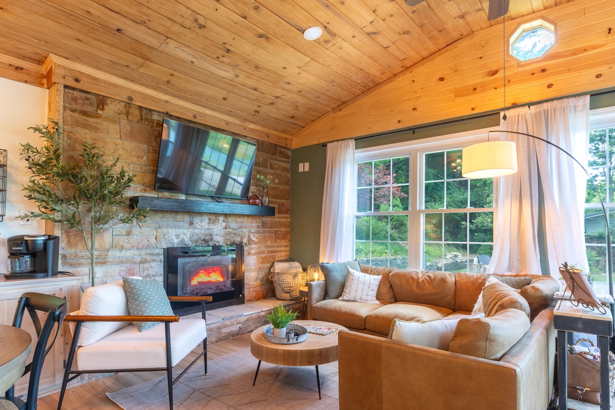 A cozy living area features a sectional sofa arranged around a circular coffee table. A stone fireplace is situated beneath a mounted television. Large windows allow natural light to fill the space, with soft curtains enhancing the serene atmosphere.