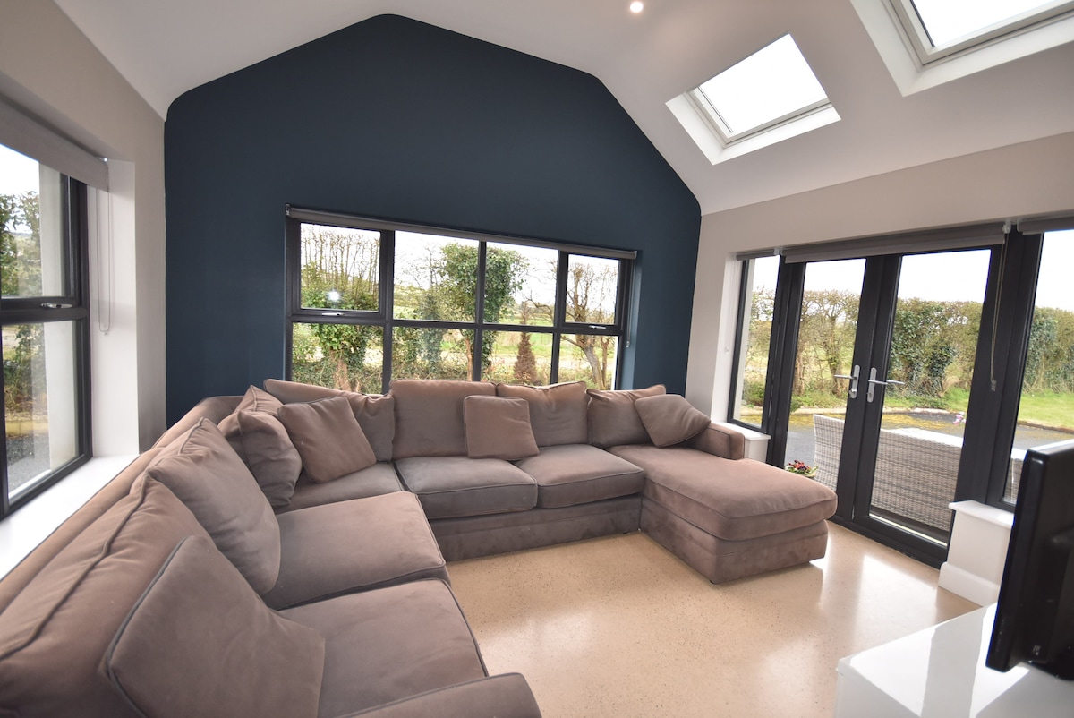A spacious living area features a large sectional sofa in neutral tones, providing ample seating. Large windows allow natural light to fill the space, with views of greenery outside. Skylights enhance brightness, and a television is positioned nearby.