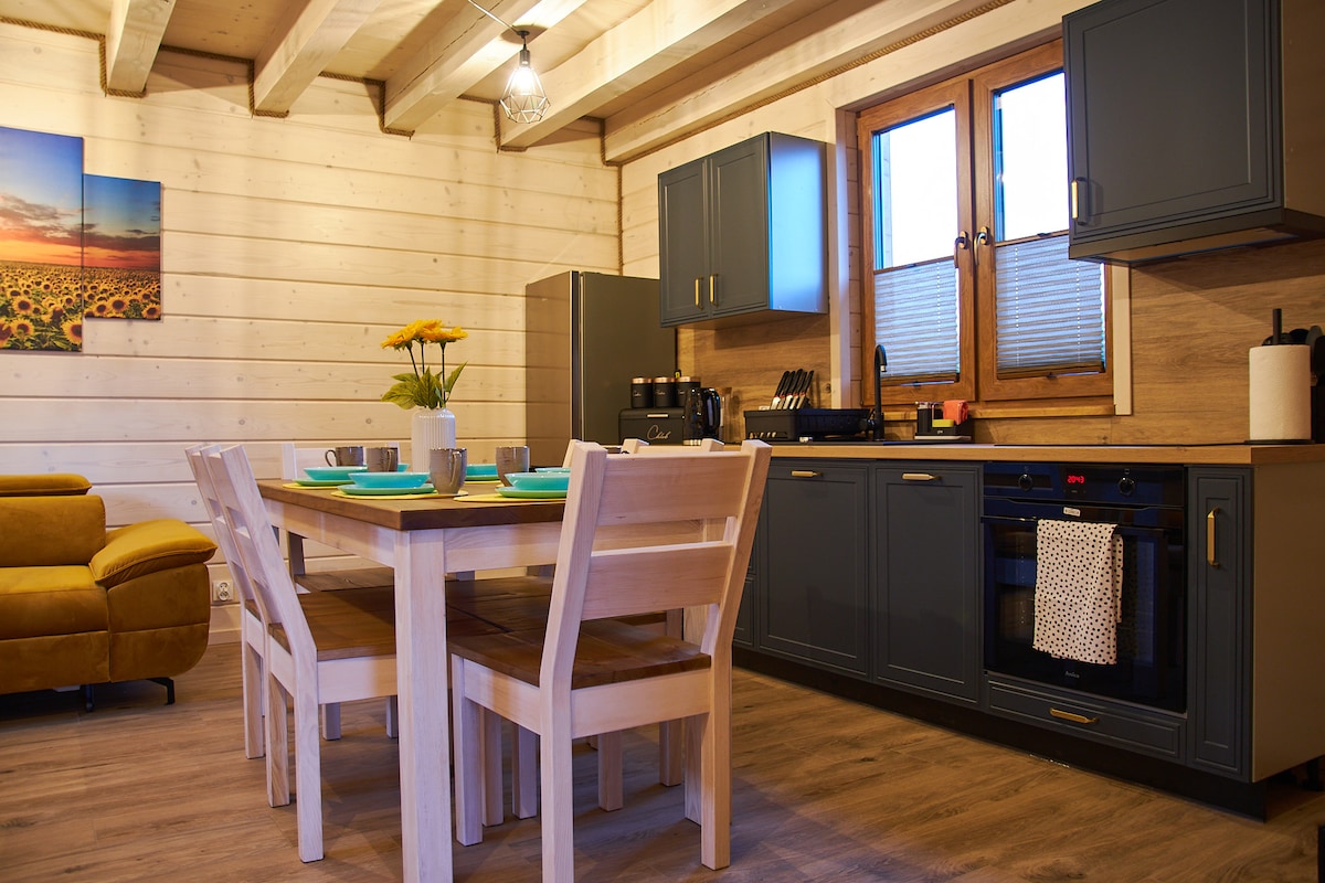 The kitchen and dining area feature modern cabinetry and a wooden dining table surrounded by four chairs. A vase of flowers sits at the center of the table. Natural light enters through a window, illuminating the space.