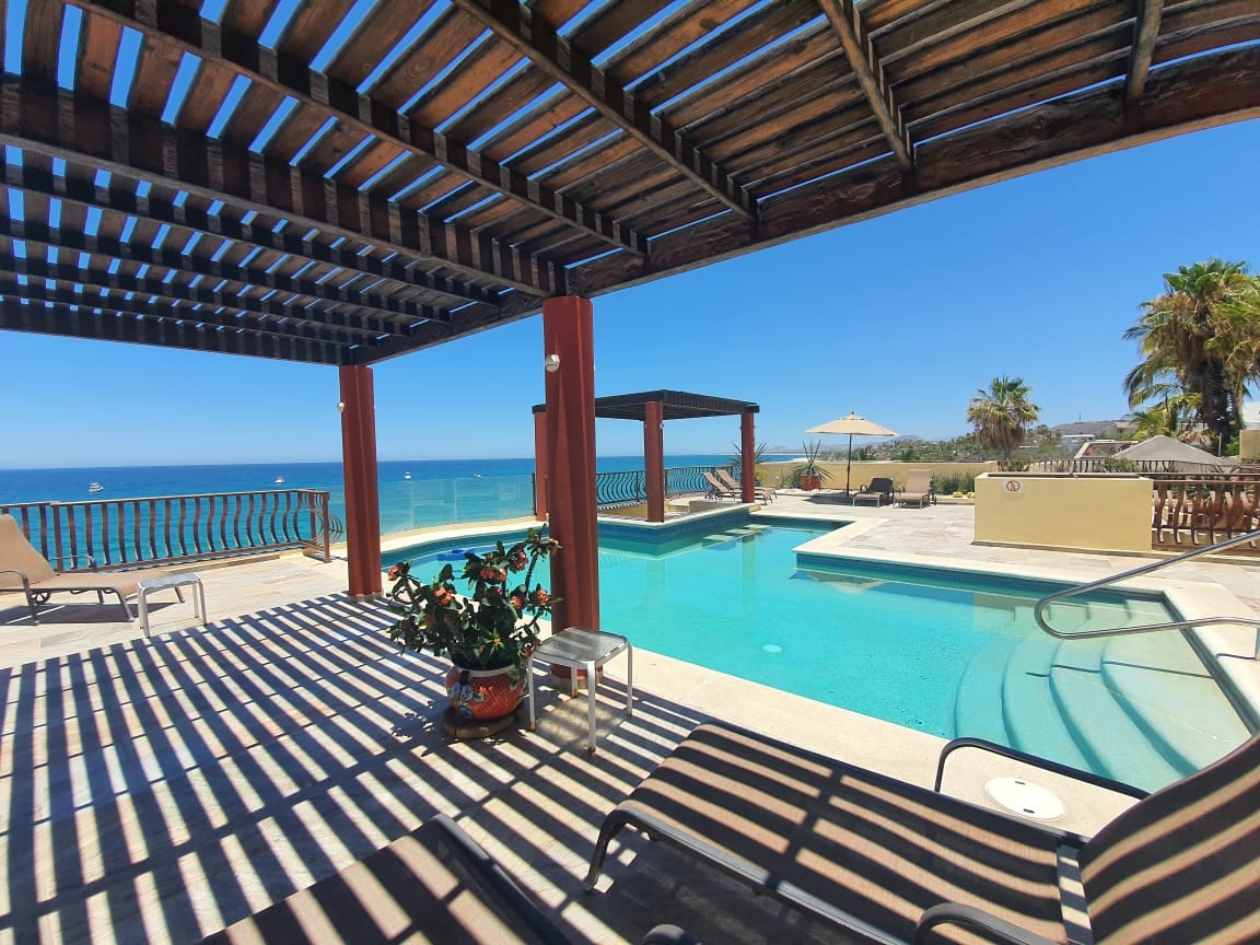 A shaded outdoor patio features a sparkling swimming pool surrounded by lounge chairs. The view overlooks a tranquil ocean, with palm trees in the background. The pergola creates interesting patterns of light and shadow on the deck.