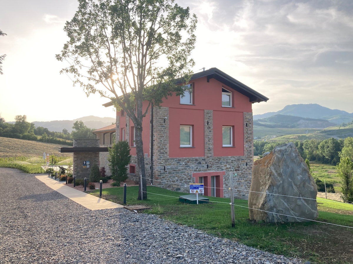 A rustic building features a combination of stone and pink exterior walls, set against a backdrop of rolling hills. Tall trees provide shade near the entrance, while a large rock adds a natural element to the landscape. The path is lined with gravel and greenery.
