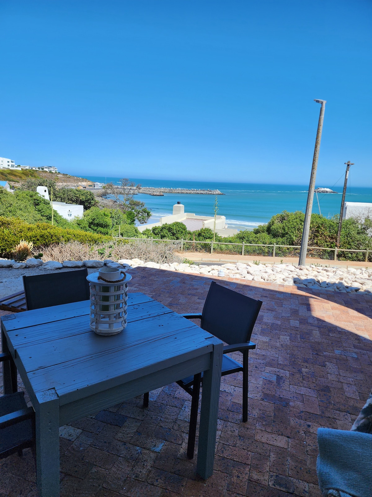 A dining table with four chairs is positioned on a patio, offering views of the ocean and the distant harbor. The blue sky above complements the natural greenery surrounding the area, creating a serene outdoor space ideal for relaxation.