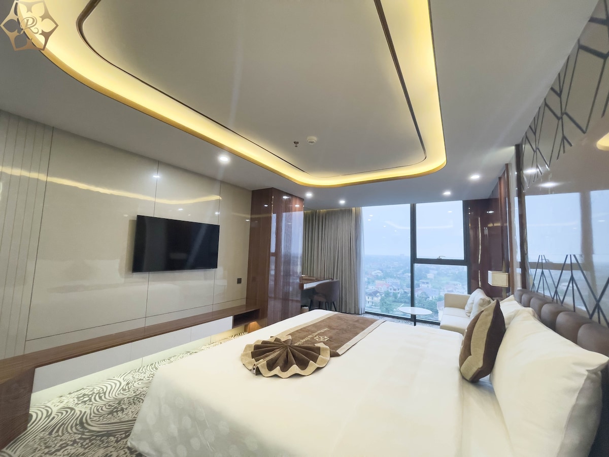 A spacious bedroom features a large bed with a soft white duvet and decorative pillows. A flat-screen television is mounted on the wall opposite the bed. Large windows provide views of the surrounding landscape, while modern lighting enhances the elegant design.