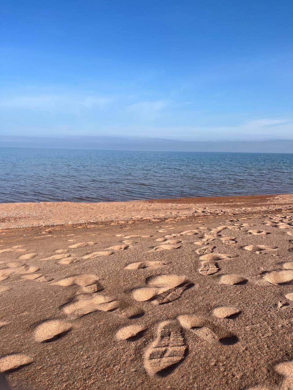 A sandy beach stretches towards calm waters under a clear blue sky. Footprints mark the smooth surface of the sand, leading towards the tranquil lake. The gentle waves ripple softly in the background, creating a serene atmosphere.