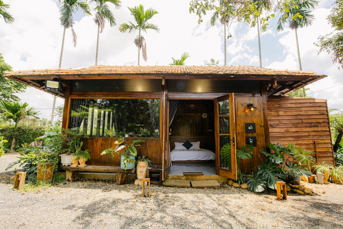 A charming wooden bungalow is surrounded by lush greenery, featuring large glass windows and doors that open to the outdoors. Potted plants adorn the entrance, while tall palm trees provide shade. The inviting exterior blends harmoniously with the natural landscape.